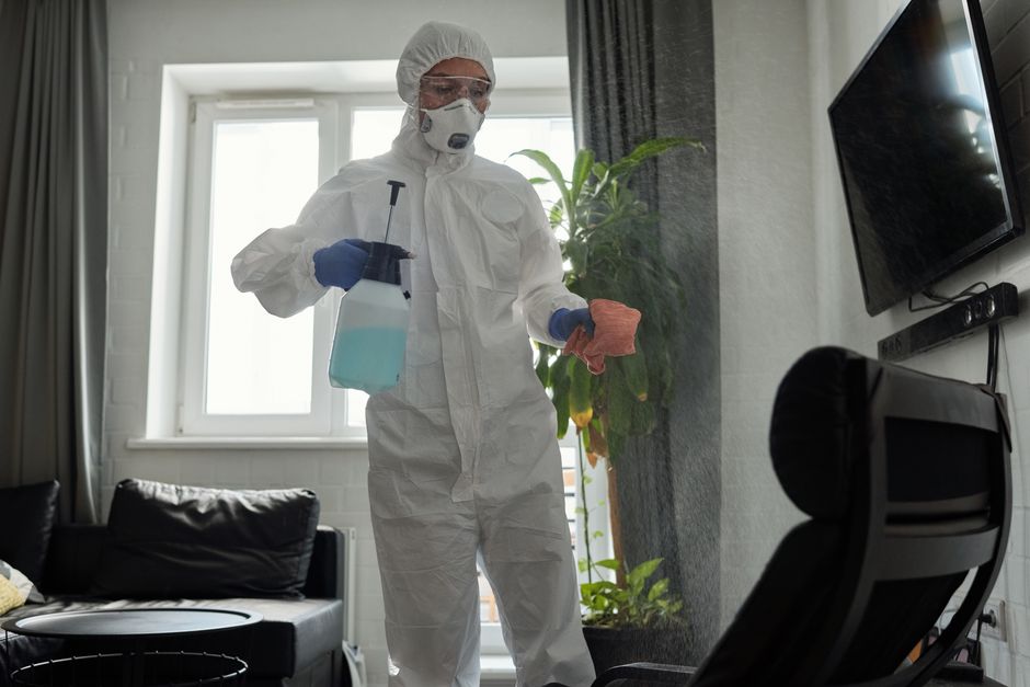 A professional cleaner dressed in protective gear sanitizes an indoor area with a spray bottle and cloth.